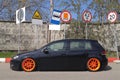 Black hatchback with orange wide wheels tuned by air suspencion parked under different road signs