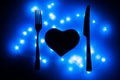 Black heart, fork and knife in the dark surrounded by blue garlands. The heart is served for dinner. Table setting for Valentine`
