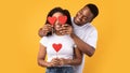 Black Husband Covering Wife`s Eyes With Hearts Over Yellow Background