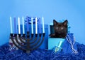 Black kitten peaking out of a blue present box next to menorah with dreidel for Hanukkah