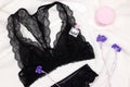 Black lace lingerie set of bralette and pantie with perfume and face cream on white fur, top view. Women essentials