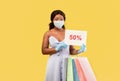 Black lady in medical mask and rubber gloves holding shopping bags and FIFTY PERCENT OFF sign, offering huge sale