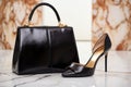 a black leather handbag next to golden high heels on a marble floor