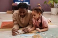 Black little girl solving jigsaw puzzle laying on floor with father