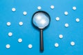 Black magnifying glass is on blue background surrounded by white pills as ornament polka dots. Medical or pharmaceutical concept b