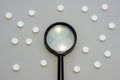 Black magnifying glass is on grey background surrounded by white pills as ornament polka dots. Medical or pharmaceutical concept b