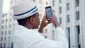 Black man tourist in white suit and glasses holds smartphone and films highrise megalopolis office buildings