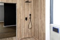 Black, matt metal concealed shower faucet and large rain shower protruding from the tiled wall, imitating wood.