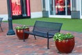 A black metal bench on a red brick sidewalk between to round brown flower pots filled with colorful flowers with lush green leaves Royalty Free Stock Photo