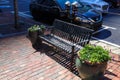 A black metal bench with two round brown flower pots filed with colorful flowers with lush green leaves on a red brick sidewalk Royalty Free Stock Photo