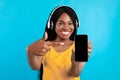 Black Millennial Woman Showing Phone Screen Wearing Headphones, Blue Background