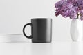 Black mug mockup with a book and a bouquet of lilac in a vase on a white table Royalty Free Stock Photo