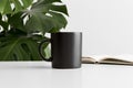 Black mug mockup with a notebook on a white table and a monstera plant