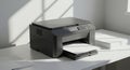 A black multifunction printer sits on a white desk, positioned near a window