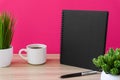 Black notebook with coffee cup and potted plant on wood desk with pink background.