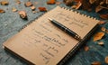 black pen rests on brown notepad with handwritten text, accompanied by scattered autumn leaves on blue Royalty Free Stock Photo
