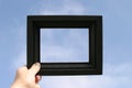 Black picture frame is held against a blue sky by a real human hand