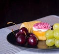 A black plate with cherries, grapes, a banana, a cupcake lies on a table in the background