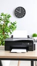 Black printer printing a blank white document on a wooden desk near a clock and plants Royalty Free Stock Photo