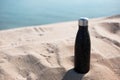 Black reusable steel stainless thermo water bottle in sand on the beach.