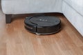 black robotic vacuum cleaner runs under sofa in room on laminate floor modern smart cleaning technology housekeeping Royalty Free Stock Photo