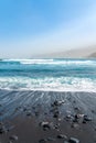 Black sand beach with rocks. Oceanic waves crashing. Blue sky and mountains in a fog. Tenerife, Spain
