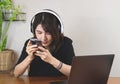 Black shirt wearing headphones, sitting at wooden table with computer laptop, using mobile phone. white wall with house plant Royalty Free Stock Photo