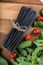 Black squid ink tinted tagliatelle pasta, uncooked, with basil and tomatoes in the background, on a wooden board Royalty Free Stock Photo