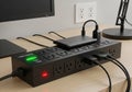 Black surge protector on a wooden desk, with multiple devices plugged in. A green \