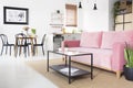 Black table in front of pink sofa in bright flat interior with poster above chairs near kitchenette. Real photo