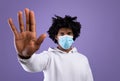 Black teen guy in face mask gesturing STOP on violet studio background