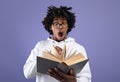 Black teen student looking at textbook in shock, afraid of too much homework on violet studio background