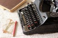 Black vintage metal type writer next to a pile of old note books, pair of eyeglasses and a pencil on a linen tablecloth