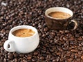 Black and white cups with freshly brewed espresso stand in coffee beans. Coffee grains are scattered on a wooden table. Close-up.