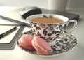 A black and white floral tea cup with pink French macaroons on a table with laptop computer and a mouse - a work from home workst