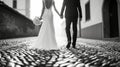 Black and white image of bride and groom holding hands and walking on a cobblestone street, capturing a moment of Royalty Free Stock Photo