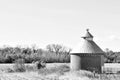 Black and white image of an old round grain silo in the countryside near a farm.