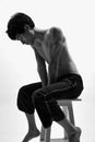 Black and white image of young handsome man posing shirtless in black trousers on high chair against white studio Royalty Free Stock Photo