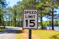 A black and white 15 MPH sign surrounded by lush green trees and plants, park benches and a lake with blue sky Royalty Free Stock Photo