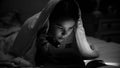 Black and white photo of girl lying under blanket and reading book with flashlight Royalty Free Stock Photo