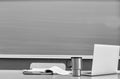 Black and white photo of laptop on professor desk with tumbler and book against black board in classroom