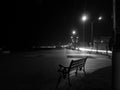 A black and white photo of a night, an empty bench at the beach in the district of Atakum, SAMSUN, and the lights of the shore. Royalty Free Stock Photo