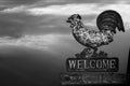 Black and white photo of an old rusty metal sign with forged rooster figurine and the inscription Welcome against stormy sky with