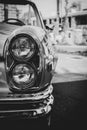 A black and white portrait of the front right headlights of an old timer car in the back the steering wheel is visible but blurred