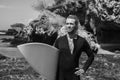 Black and white portrait of handsome man surfer in black wetsuit, holding white surf board  and blue ocean, cliff, rocks behind Royalty Free Stock Photo