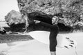 Black and white portrait of handsome man surfer in black wetsuit, holding white surf board  and blue ocean, cliff, rocks behind Royalty Free Stock Photo