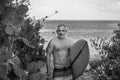 Black and white portrait of handsome shirtless man surfer, holding white surf board and cactus on background