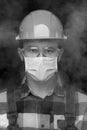 Black and white portrait of a worker in a helmet, mask and safety glasses amid smoke.