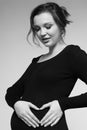 Black and white Portrait of young pregnant female in black dress with hands near pregnant belly with heart sign