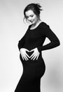 Black and white portrait of pregnant female in black dress with hands near pregnant belly with heart sign